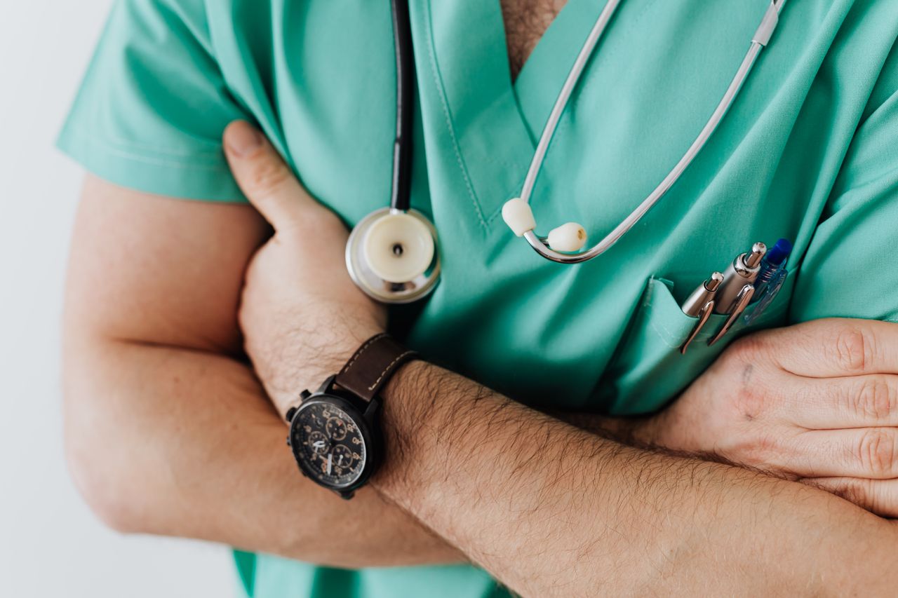 Unrecognizable man wearing medical uniform with stethoscope and pens standing in hospital with crossed arms during work in day time